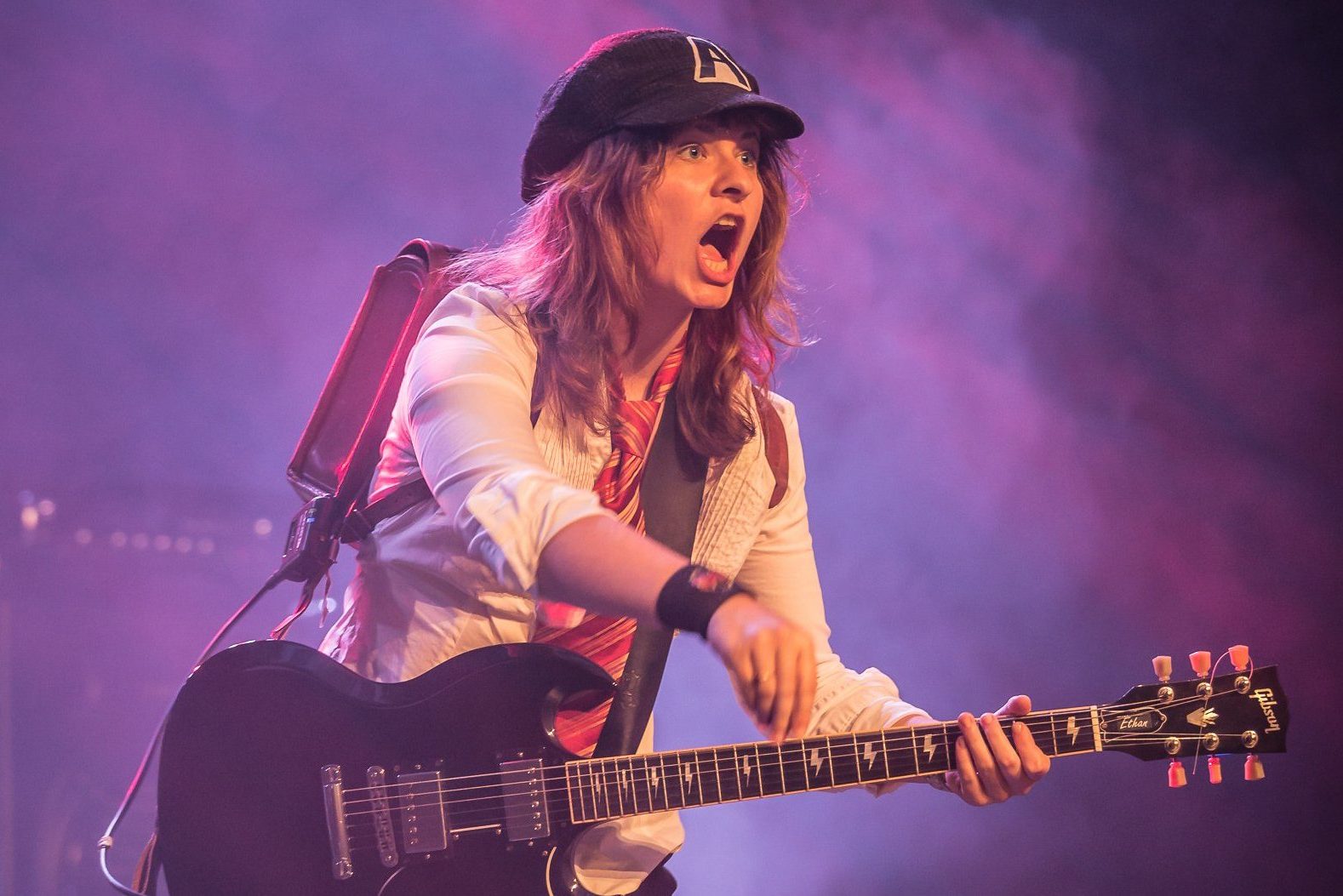 Weibliches Angus-Young-Double auf der Bühne mit Schulranzen und weißer Bluse, lila Hintergrund