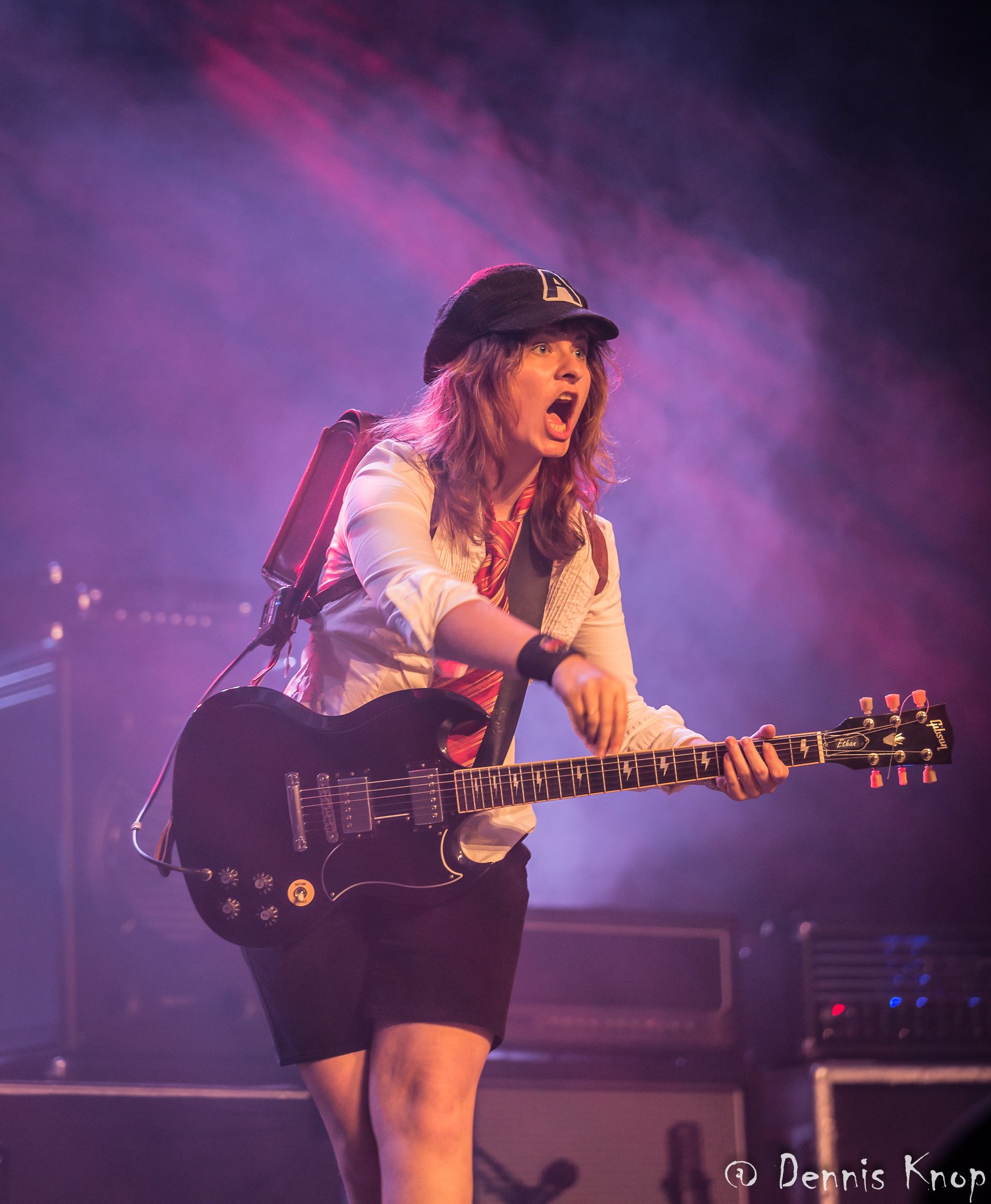 Weibliches Angus-Young-Double auf der Bühne mit Schulranzen und weißer Bluse, lila Hintergrund