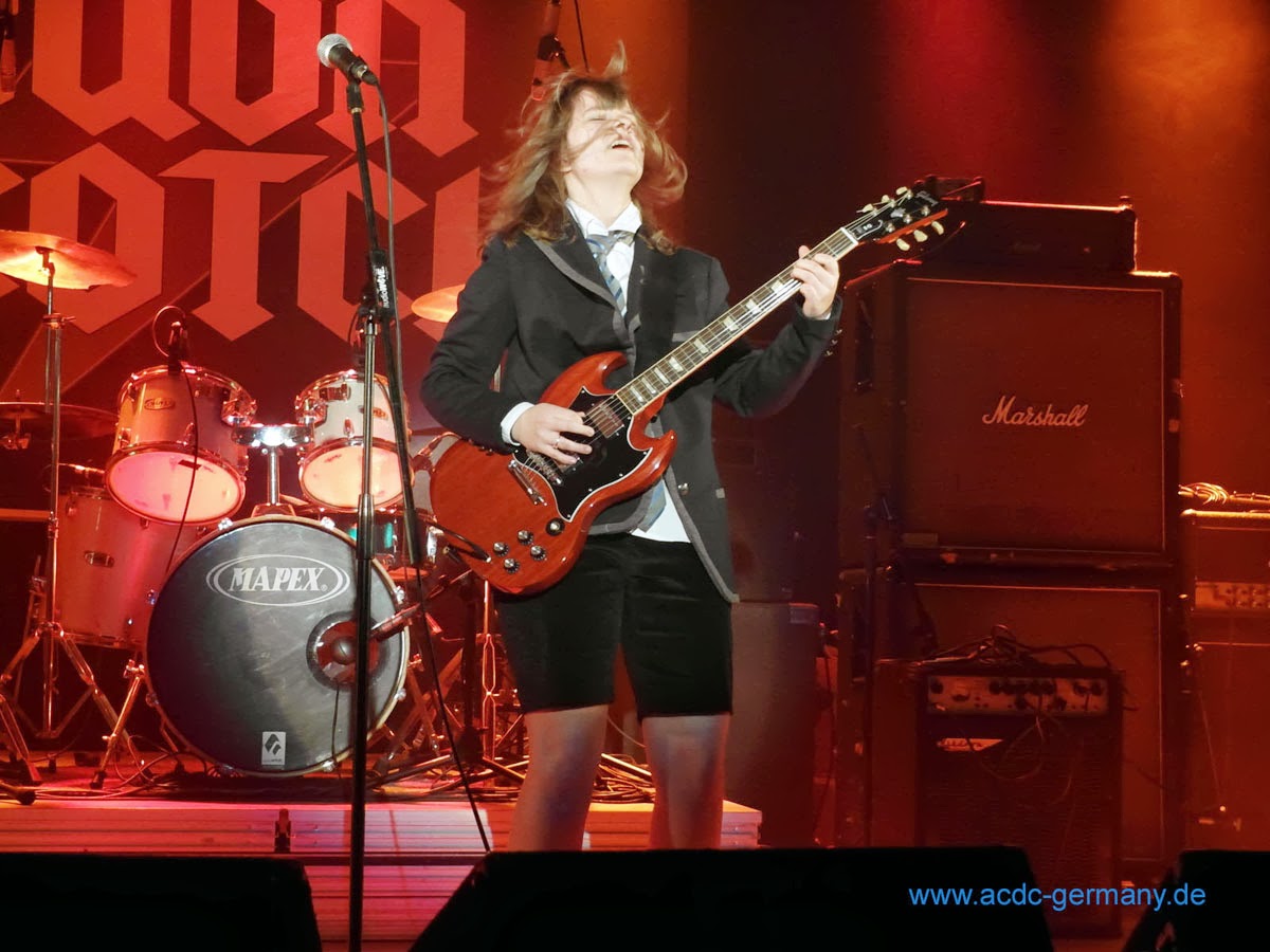 Weibliches Angus-Young-Double in Aktion on stage, rote E-Gitarre, rotes Bühnenlicht, Schlagzeug, Amp