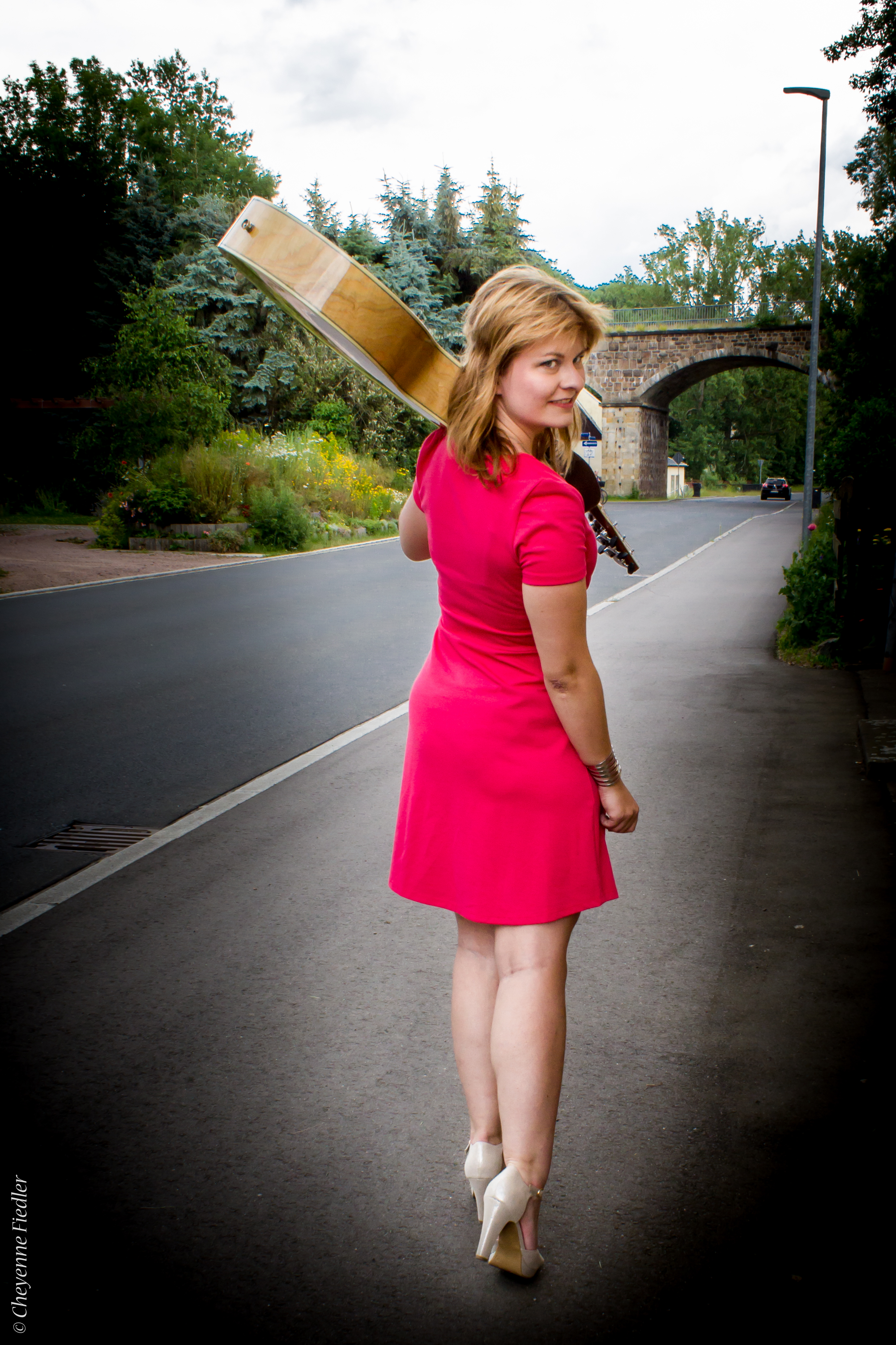 Laufende Frau in rosa Kleid von hinten, Kopf zur Kamera gedreht, Akustikgitarre auf der Schulter