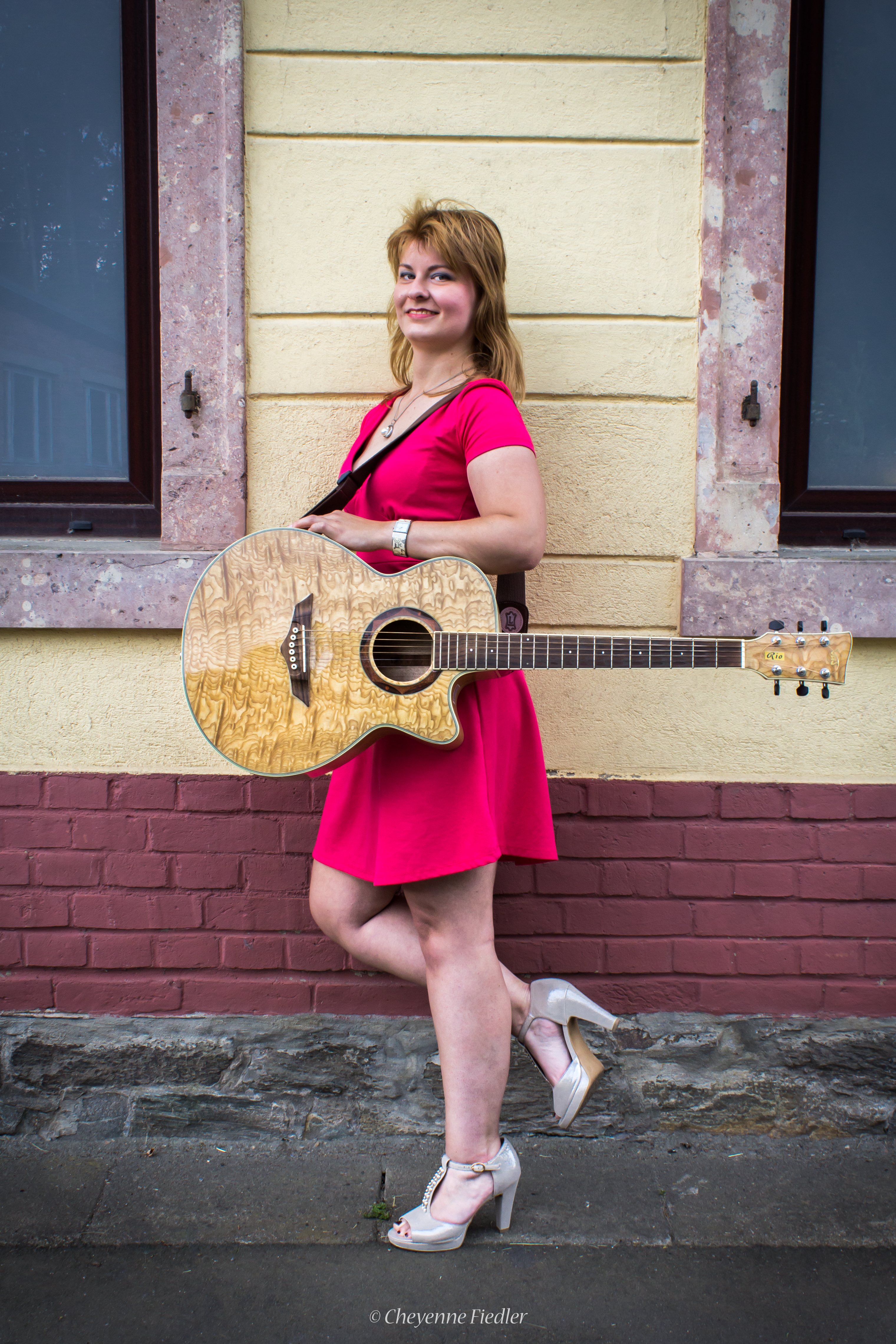 Stehende Frau mit rosa Kleid, rechtes Bein angewinkelt, schaut in die Kamera, mit Akustikgitarre