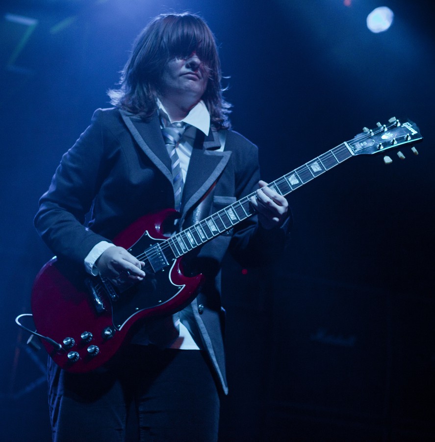 Weibliches Angus-Young-Double spielt auf der Bühne, mit E-Gitarre, dunkelblaues Bühnenlicht