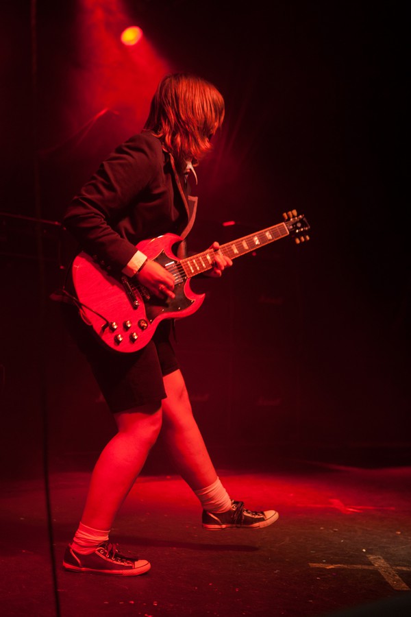 Weibliches Angus-Young-Double läuft und spielt auf der Bühne, rotes Licht, seitliche Perspektive