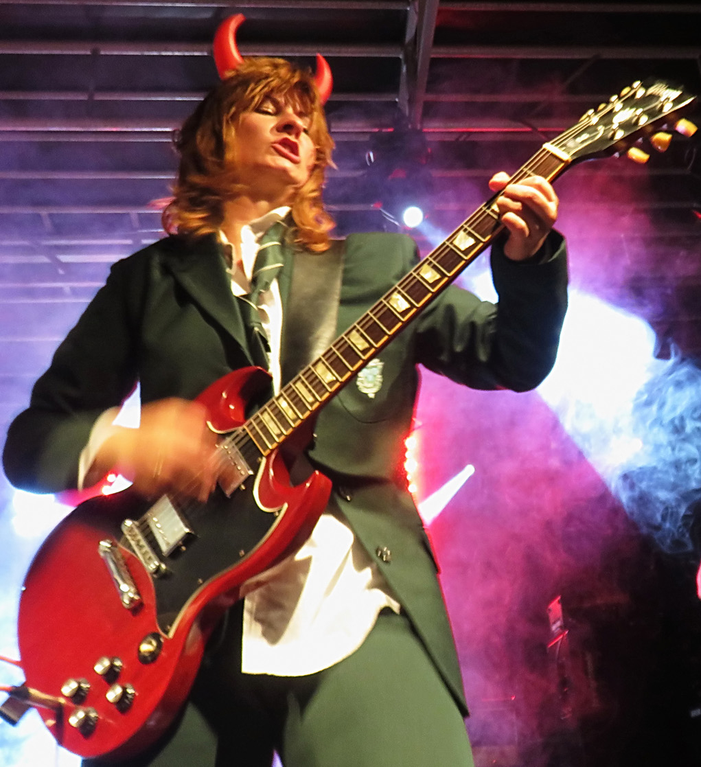 Weibliches Angus-Young-Double mit roten Hörnern und E-Gitarre spielt auf der Bühne, lila Hintergrund