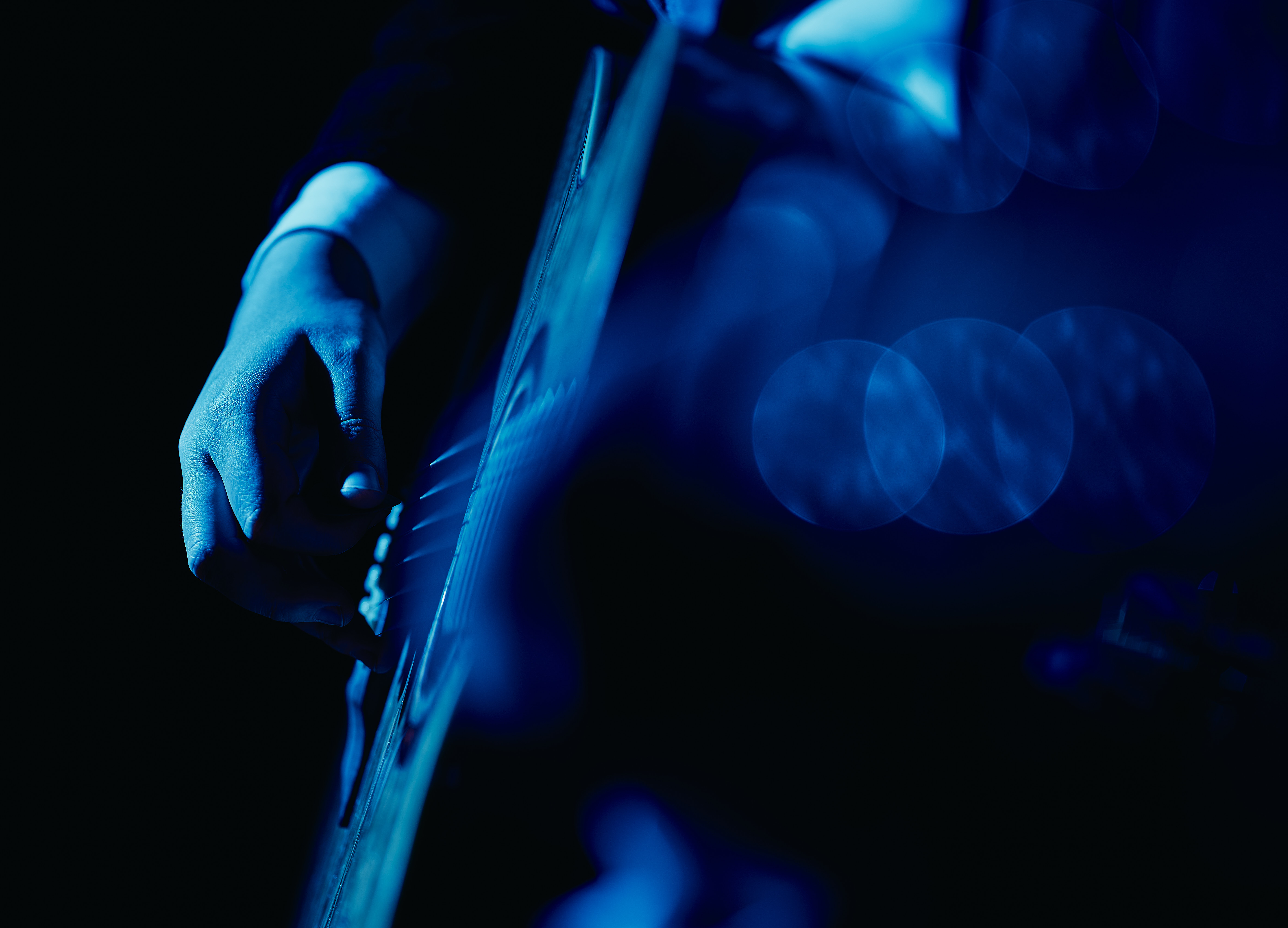 blau-schwarze Nachaufnahme von der Seite eines spielenden Gitarristen, Fokus auf Hand und Korpus