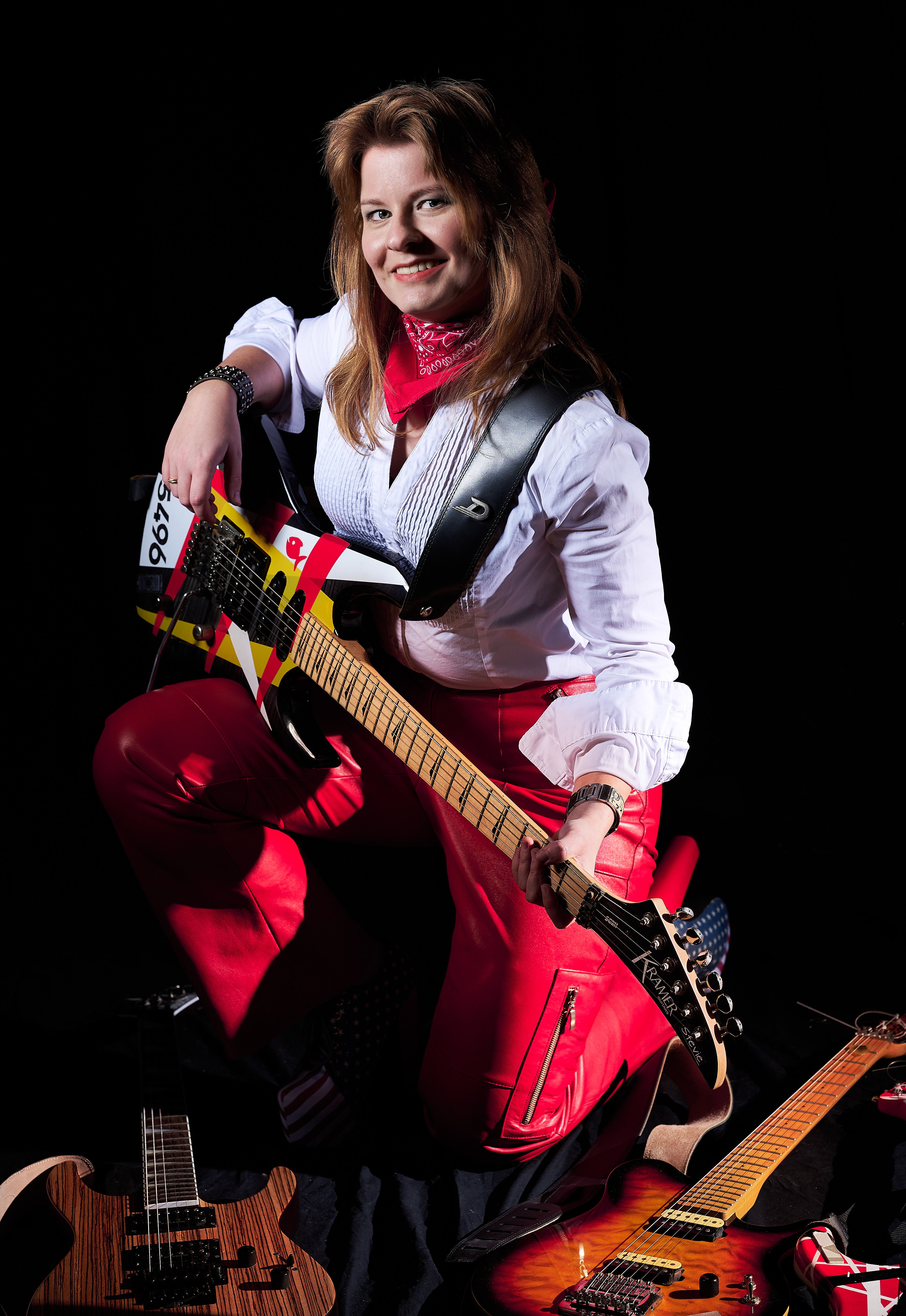Lächelnde Frau kniet in roter Lederhose, weiße Bluse, E-Gitarre in der Hand, vor ihr liegen Gitarren