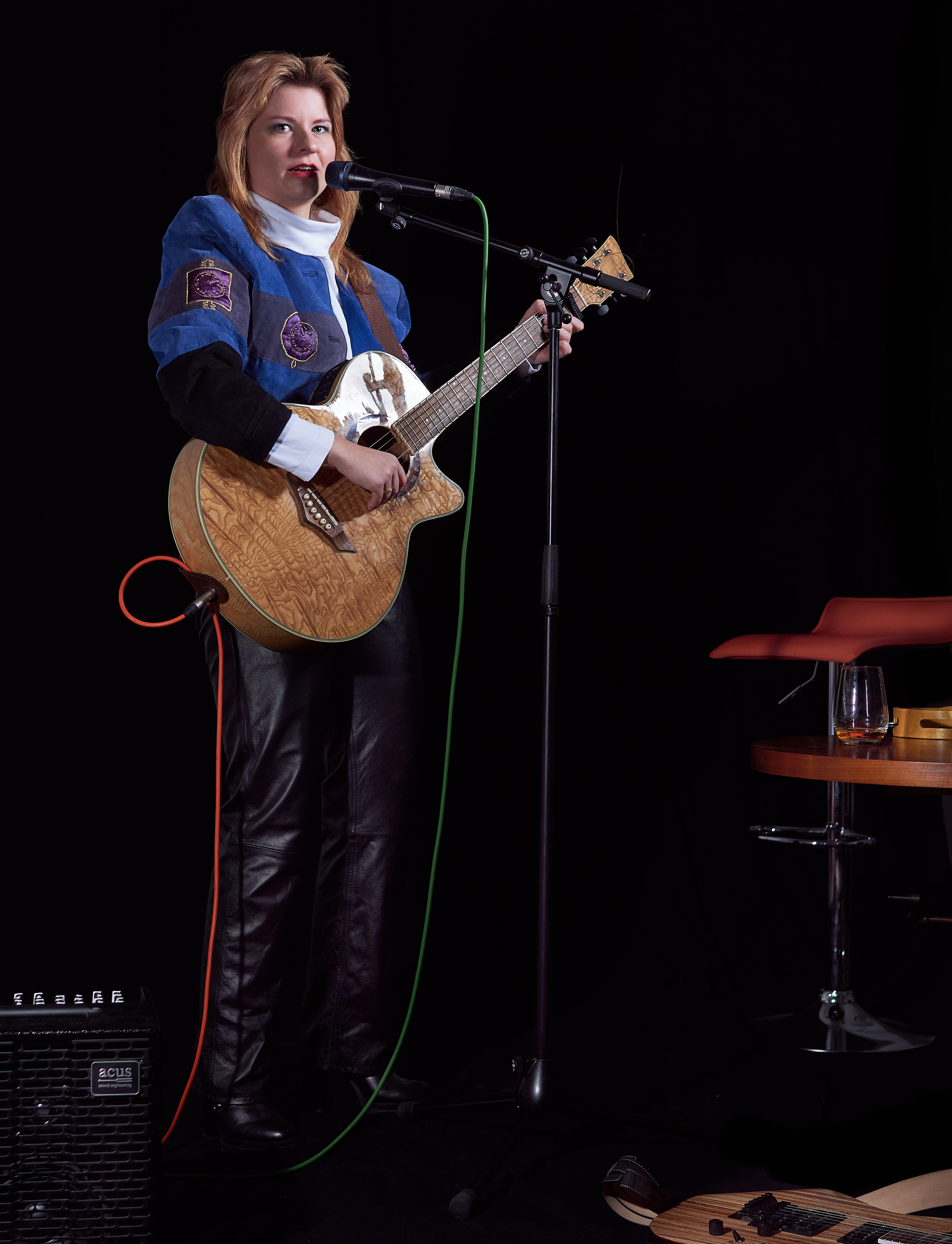 Musikerin singend mit Akustikgitarre, blauer Jakce, links steht ein Verstärker, rechts ein Barhocker