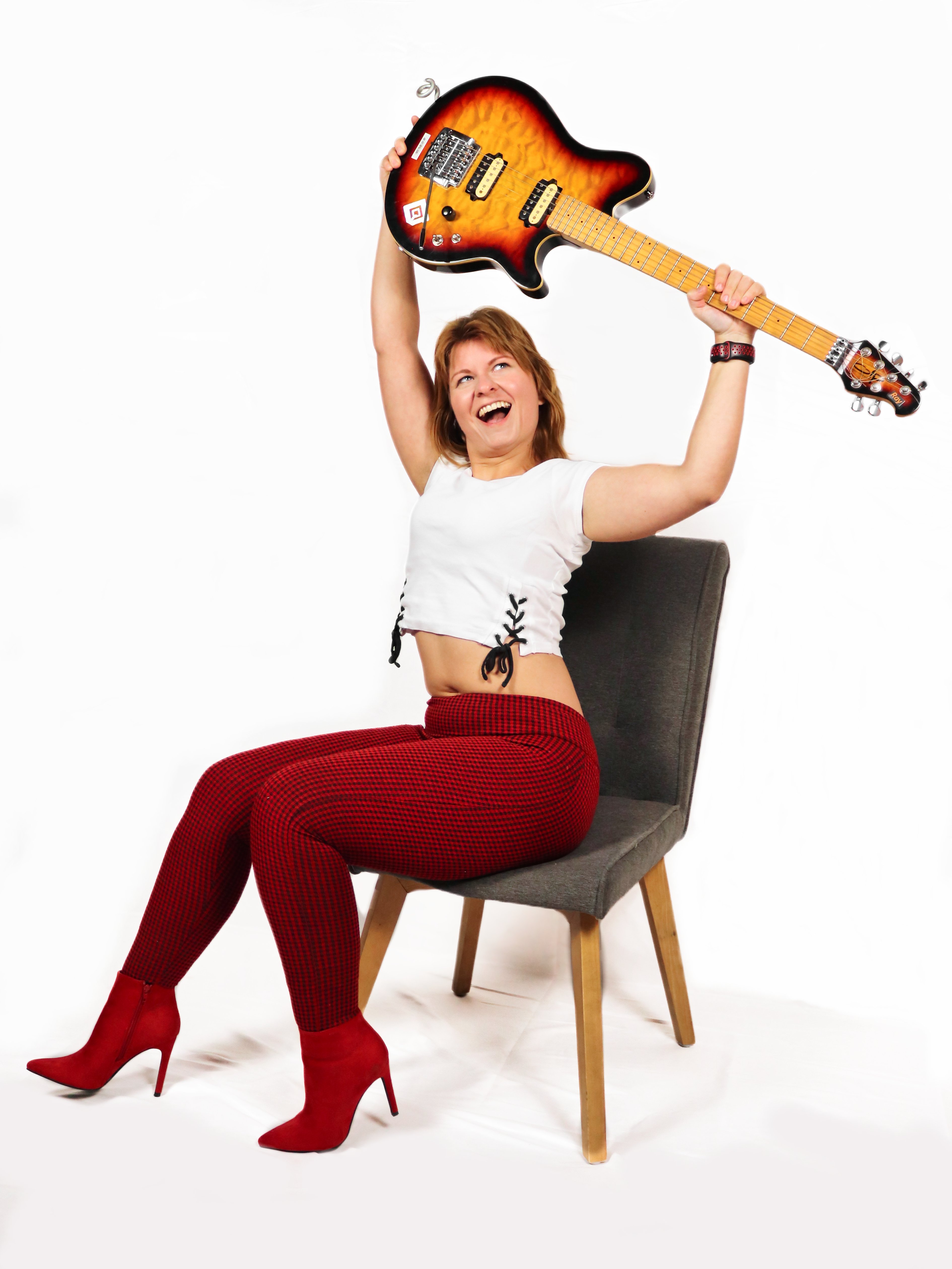 Frau sitzend in weißem Shirt, hält E-Gitarre in Sunburst Look mit energischer Pose nach oben