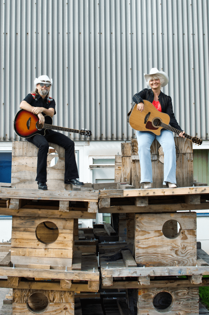 Two people with acoustic guitars sitting on a stack of wooden pallets in front of a gray metal wall; one wearing a white cowboy hat and black jacket, the other in black clothing with red details.