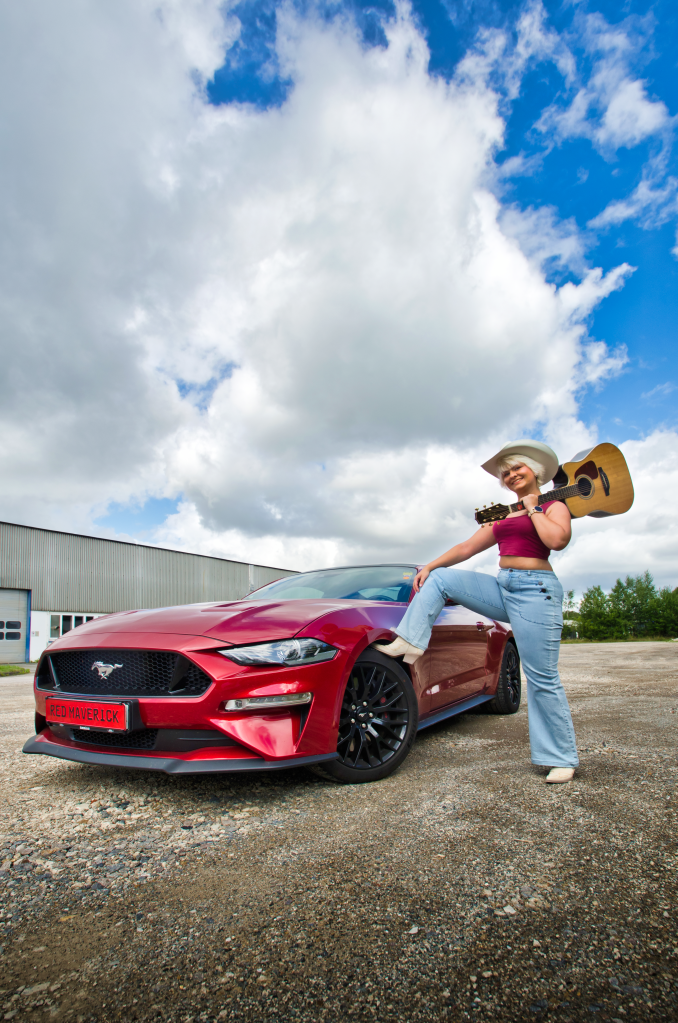 A person wearing a white cowboy hat and holding an acoustic guitar stands beside a red Ford Mustang with the “RED MAVERICK” license plate, resting one leg on the fender, with a cloudy sky and an industrial building in the background.