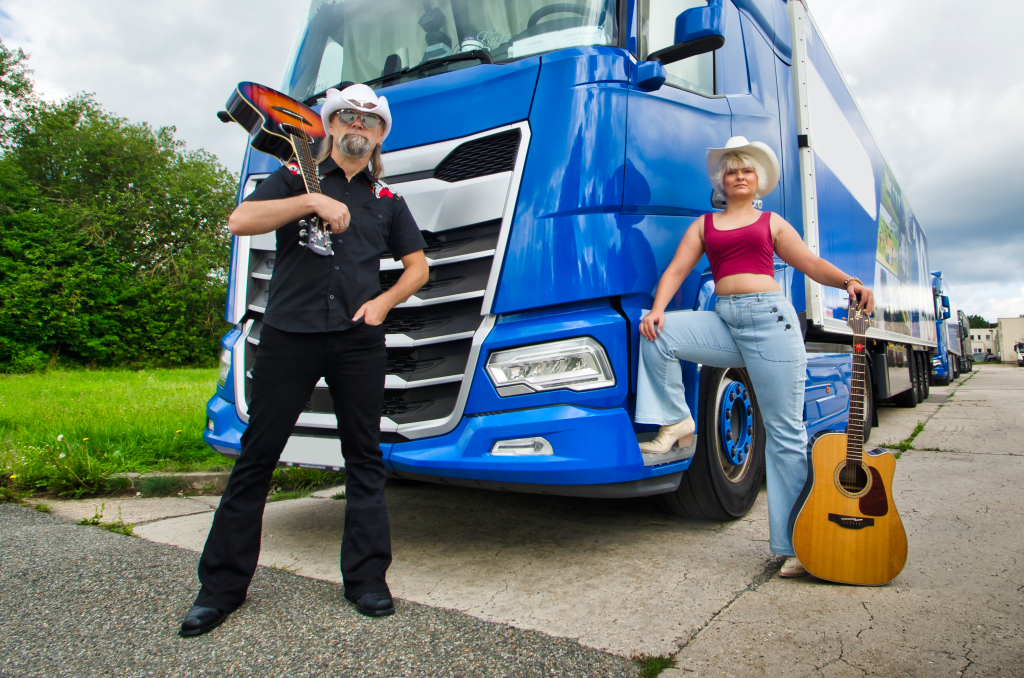 Two members of the Hood Up band project holding guitars in front of a large blue truck on a road.