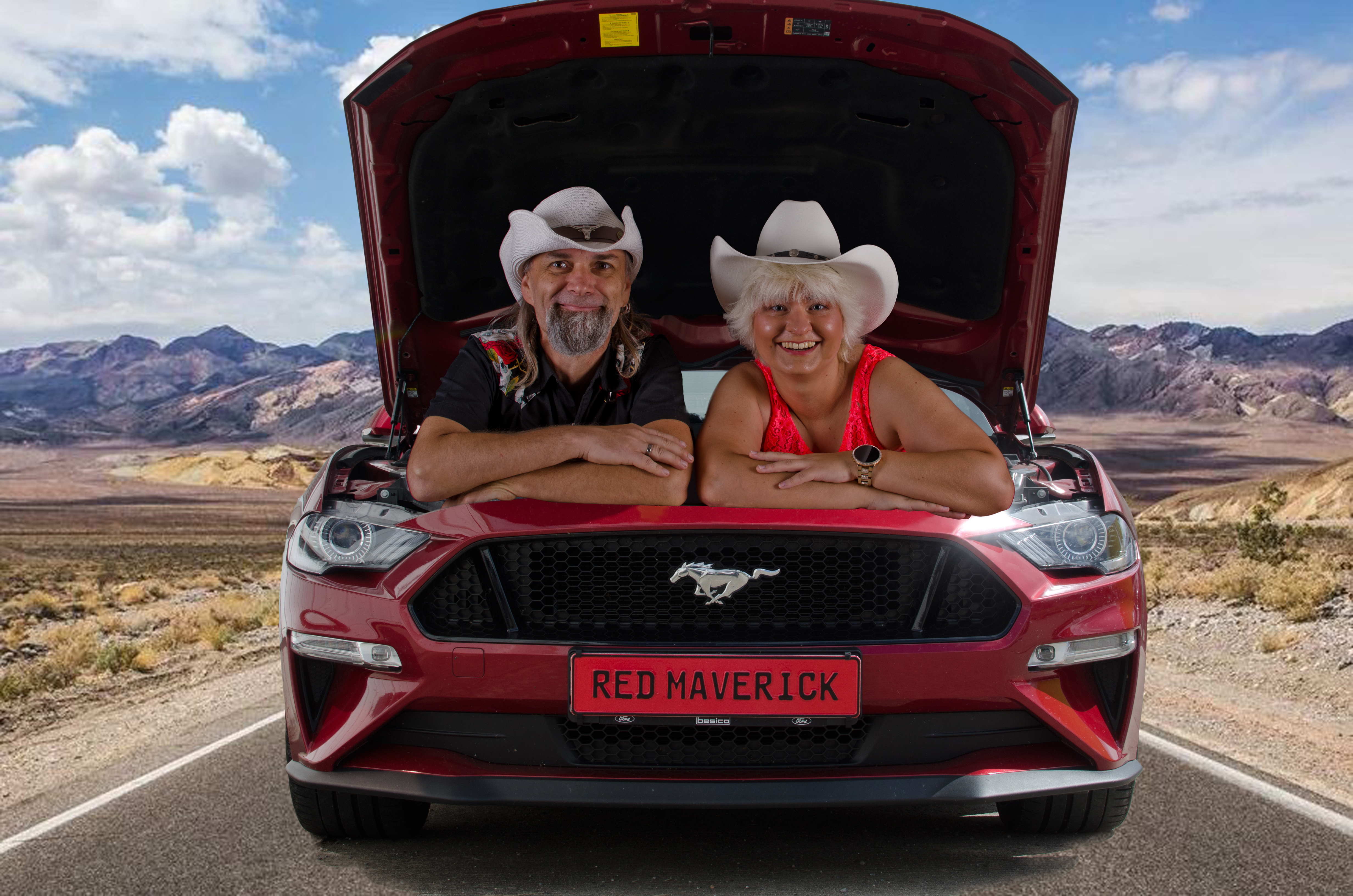 Two people wearing cowboy hats are smiling as they lean over the open hood of a red Ford Mustang, parked on a deserted road with a mountainous desert landscape in the background.
